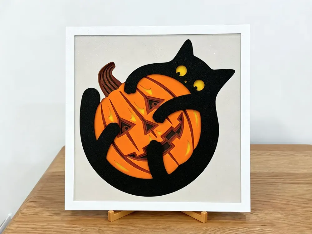 Halloween layered art: a black cat with yellow eyes hugging a carved orange pumpkin on a cream panel, in a white frame on a stand
