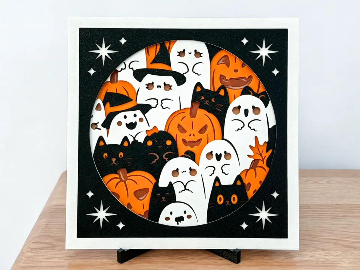 Halloween layered art: pumpkins, ghosts and black cats in a round window, with a white border and a black panel with cut-out stars, on a stand